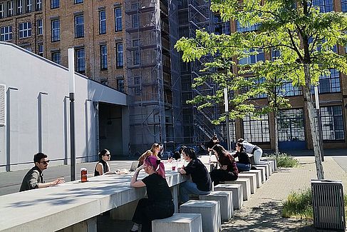 Mehrere Personen sitzen beidseitig entlang einer langen Steintafel mit quadratischen Klötzen als Sitzgelegenheit. Sie sind zueinander gewandt und ins Gespräch vertieft. © HTW Berlin / Lisa Pichler