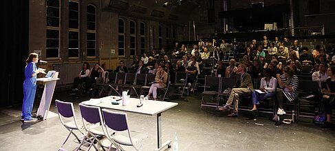 Wortbeitrag auf der Bühne mit aufmerksamem Publikum (Tag 1). © IKTf Berlin / Janosch Weiss